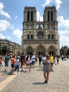 Mulher com macaquinho listrado azul e branco e com flores rosas usando óculos de sol na frente na catedral Notre Dame. Diversas outras pessoas estão em uma fila e aparecem na foto