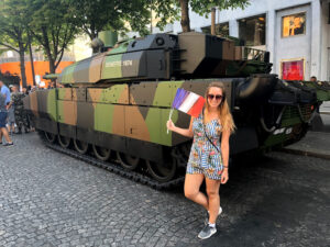 Mulher loria vestida com um macaquinho listrado azul e branco com flores na frente de um tanque de guerra segurando uma bandeira da frança