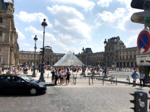 Louvre visto pelo lado de fora - Pirâmide de vidro bem ao centro e muitas pessoas espalhadas pela foto